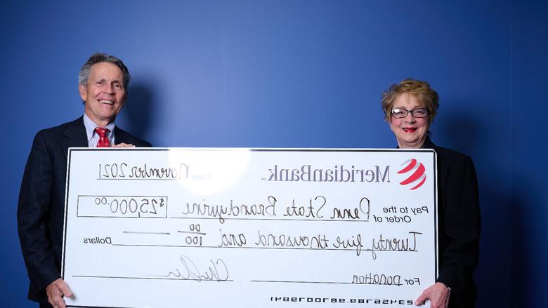 Meridian CEO Chris Annas with 宾州州立大学白兰地酒 总理 Marilyn 井 stand in front of a blue wall holding a large white check. 