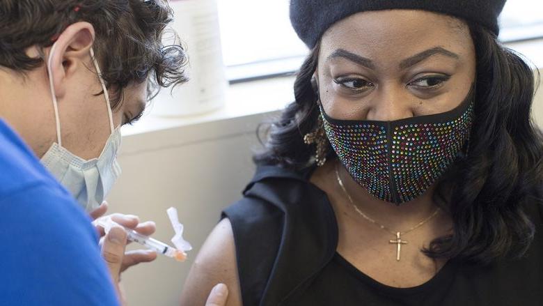 Tamika华盛顿, 谁戴着珠宝面具, 十字架项链和贝雷帽, 当医护人员准备插入装满新型冠状病毒肺炎疫苗的注射器时，她把目光从裸露的手臂上移开.